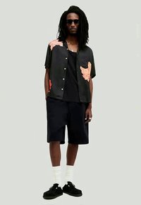 Muž s dlouhými dredy a slunečními brýlemi má na sobě černou košili s krátkým rukávem s oranžovými potisky listů, černé kraťasy, bílé ponožky a černé boty.