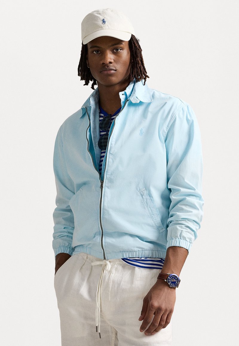 Hellblaues Zip-Jacket mit elastischen Bündchen und Seitentaschen, getragen über einem gestreiften Hemd. Kombiniert mit einer weißen Mütze und einer braunen Uhr.
