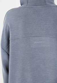 Rückansicht einer Person, die einen grauen Kapuzenpullover trägt, mit einer Naht über dem oberen Rücken und schwachem Schriftzug "ENDURANCE" unter der Naht.