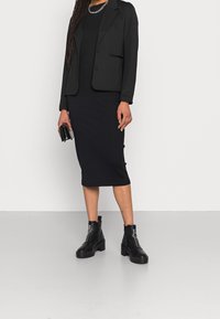 Blazer noir porté sur une robe noire côtelée, associé à des bottines noires épaisses. Le blazer présente deux poches avant et une coupe ajustée.