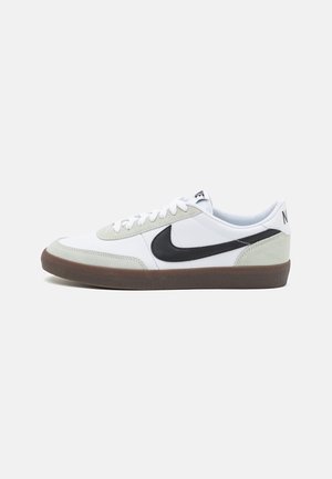 Białe niskie buty sportowe z jasnoszarymi zamszowymi akcentami, czarnym logo Nike, białymi sznurowadłami i brązową gumową podeszwą na jasnym tle.