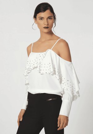 Mujer con cabello oscuro lleva una blusa blanca con hombros descubiertos, volantes y tachuelas plateadas, combinada con pantalones negros sobre un fondo claro y liso.