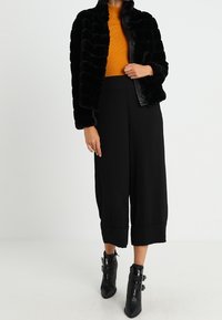 Veste en fausse fourrure noire avec des rayures horizontales, pull côtelé orange, pantalon large noir et bottines avec des accents de boucle.