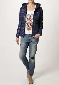 Granatowa kurtka puchowa z kapturem, zestawiona z białym T-shirtem z kolorowym, kwiatowym wzorem, postrzępionymi niebieskimi dżinsami oraz beżowymi botkami za kostkę.