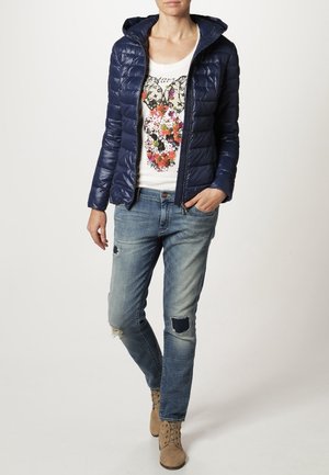 Laivastonsininen toppatakki hupulla, yhdistettynä valkoiseen graafiseen T-paitaan, jossa on värikäs kukkakuosi, kuluneet siniset farkut ja vaaleanruskeat nilkkurit.