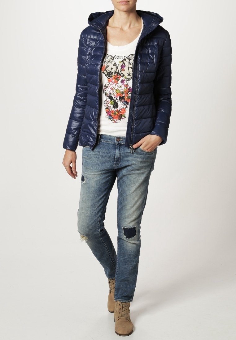 Granatowa kurtka puchowa z kapturem, zestawiona z białym T-shirtem z kolorowym, kwiatowym wzorem, postrzępionymi niebieskimi dżinsami oraz beżowymi botkami za kostkę.