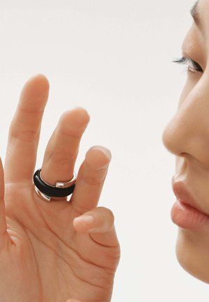Anillo negro y plateado con una superficie lisa, que presenta un diseño moderno y formas superpuestas, utilizado en un dedo sobre un fondo neutro.