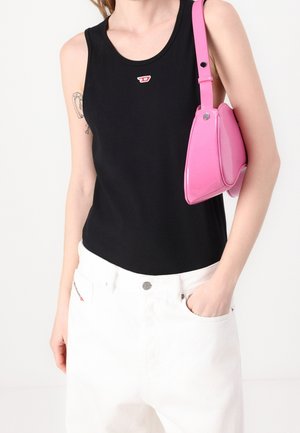 Femme portant un haut noir sans manches et un pantalon blanc, la main gauche dans la poche, portant un petit sac à bandoulière rose brillant sur le bras droit.