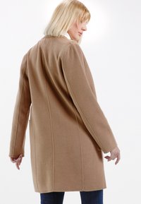 Beige Woll-Mix-Mantel mit geradem Schnitt, langen Ärmeln, Seitentaschen und sichtbaren Nähten, getragen über dunklen Jeans.