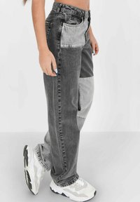 Grau- und hellgraue Patchwork-Jeans mit lockerer Passform, ausgestattet mit Vordertaschen und Reißverschluss. Kombiniert mit weißen Sneakers.