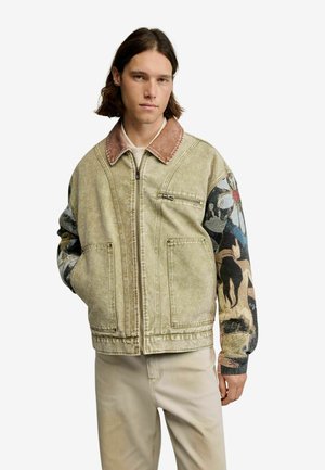 Junger Mann trägt eine beige Jacke mit braunem Kragen und künstlerisch gemusterten Ärmeln, steht mit den Händen in den Taschen vor weißem Hintergrund.