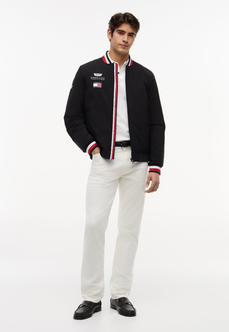 Νεαρός άνδρας όρθιος, φοράει μαύρο μπουφάν bomber με κόκκινη-λευκή λεπτομέρεια, λευκό πόλο πουκάμισο, λευκό παντελόνι και μαύρα μοκασίνια, μπροστά σε λευκό φόντο.