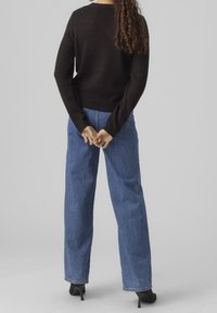 Maglione di lana nera a maniche lunghe, abbinato a jeans di denim blu a vita alta. La modella è vista di spalle, con le mani incrociate in vita.