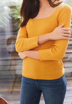 Senfgelbes Rippstrickpullover mit Dreiviertelärmeln, ausgestattet mit strukturierten Punkten und einem quadratischen Ausschnitt, kombiniert mit dunkelblauen Jeans.