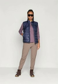 Marinblå quiltad puffer väst över en mauve långärmad topp, i kombination med ljusbruna byxor och bruna vandringskängor. Modell som bär mörka solglasögon.