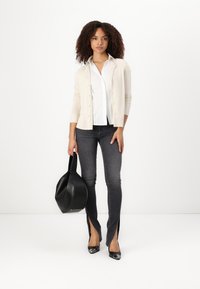 Beige Cardigan über einer weißen Bluse, schwarze Handtasche und schwarze Jeans mit seitlichen Schlitzen. Schwarze spitze Ballerinas vervollständigen das Outfit.
