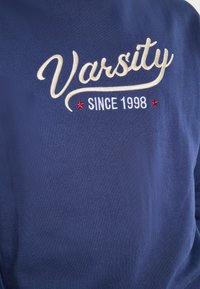 Tengerészkék pulóver, hímzett fehér "Varsity" felirattal és piros kiegészítőkkel, valamint a "SINCE 1998" felirattal fehér színben, mely hangsúlyosan a mellkason látható.