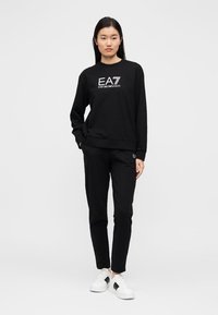 Μαύρο φούτερ με λευκό λογότυπο EA7, σε συνδυασμό με μαύρο παντελόνι. Το μοντέλο φοράει λευκά αθλητικά παπούτσια. Λείο ύφασμα, απλός σχεδιασμός, χαλαρή εφαρμογή.