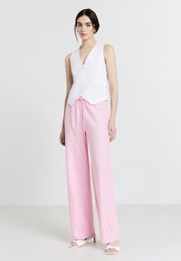 Débardeur blanc sans manches avec un motif à œillets, associé à un pantalon large rose pâle avec des coutures décoratives à l'ourlet. Sandales ouvertes blanches.