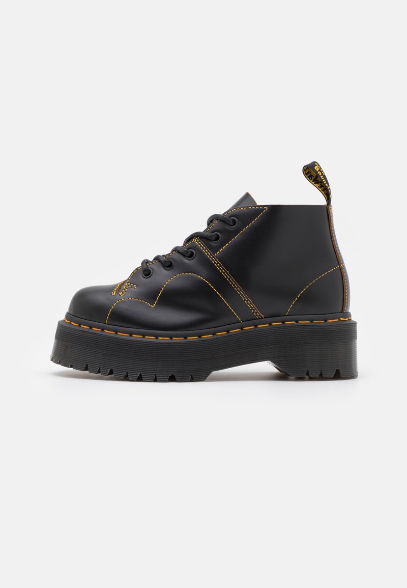 Botas negras de tobillo con suela gruesa y estriada, costuras amarillas en contraste y una lengüeta en el talón. Material de cuero suave.