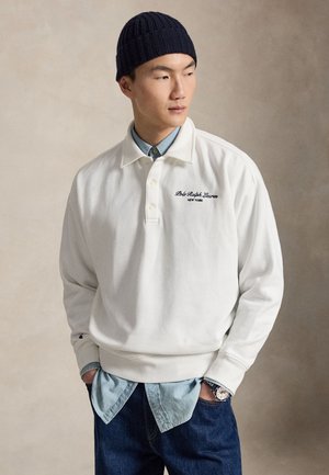 Άνδρας που φοράει άσπρη μπλούζα Polo Ralph Lauren πάνω από ανοιχτό μπλε πουκάμισο, μπλε τζιν, μαύρο πλεκτό καπέλο και ρολόι χειρός, με τα χέρια στις τσέπες.