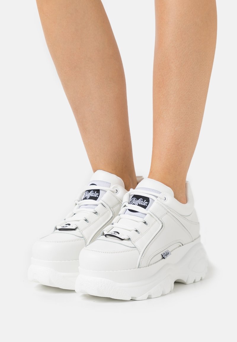 Pieds portant des baskets à plateforme blanches avec des semelles épaisses et des logos noirs "Buffalo" sur la languette et le côté, sur un fond uni.