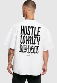 Balts, pārāk liels T-krekls ar uzrakstu uz muguras: "HUSTLE LOYALTY RESPECT" dažādos fonta stilos. Kokvilnas materiāls ar brīvu piegriezumu.
