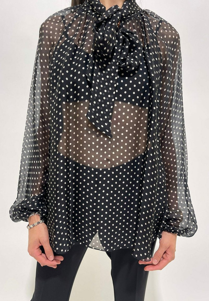 Blusa nera a pois trasparente con maniche lunghe e un fiocco legato al collo, indossata sopra intimo nero e abbinata a pantaloni neri.