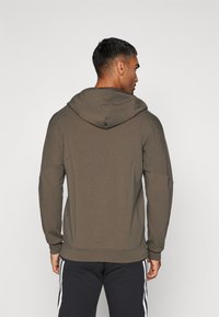 Sudadera con capucha en verde oliva, con una textura suave, mangas largas y un corte relajado. Costuras visibles y un dobladillo acanalado.