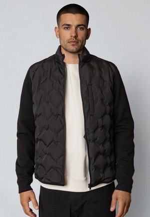 Man wearing a black quilted vest over a white sweatshirt, standing against a plain light gray background, facing forward.