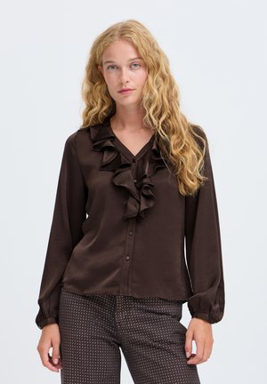 Femme aux longs cheveux blonds bouclés portant un chemisier à volants marron foncé et un pantalon marron à motifs, debout devant un fond clair uni.