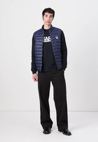 Marineblauwe puffer gilet met horizontale stiksels, gedragen over een zwart sweatshirt. Zwarte losse broek en zwarte schoenen maken de outfit compleet.
