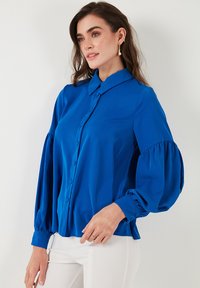 Blusa blu a maniche lunghe con chiusura a bottoni, grandi maniche a sbuffo e colletto strutturato, realizzata in tessuto liscio e leggero.