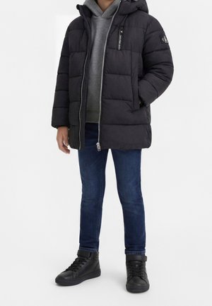 Enfant portant une doudoune noire par-dessus un sweat à capuche gris, un jean bleu et des baskets montantes noires, debout devant un fond blanc.