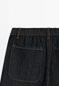 Dark denim pants featuring an elastic waistband, contrasting orange stitching, and a single back pocket with a stitched outline.