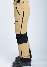 Beige og sorte skibukser med en tekstureret finish, der har lynlåslommer og forstærkede kanter. Inkluderer justerbar talje og ankelmanchetter.