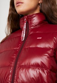 Rød polstret jakke med høj krave, glat overflade og kontrasterende hvid lynlås med brandlogo. Nærbillede af frontområdet.