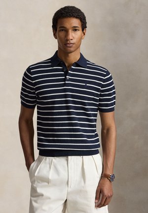Man draagt een marineblauw-wit gestreepte korte mouw polo en een witte geplooide broek met handen in de zakken.