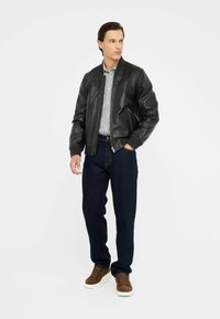 Schwarze Lederbomberjacke mit geripptem Kragen, getragen über einem hellen gestreiften Hemd, kombiniert mit dunkelblauen Jeans und braunen Wildledersneakern.