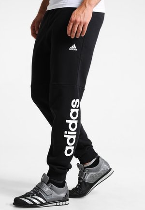 Man draagt zwarte Adidas joggingbroek met wit logo en zilvergrijze Adidas sportschoenen tegen een effen witte achtergrond.