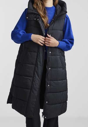 Personne portant une longue doudoune noire sans manches sur un pull bleu, en train de fermer la fermeture éclair et les boutons-pression de la doudoune.