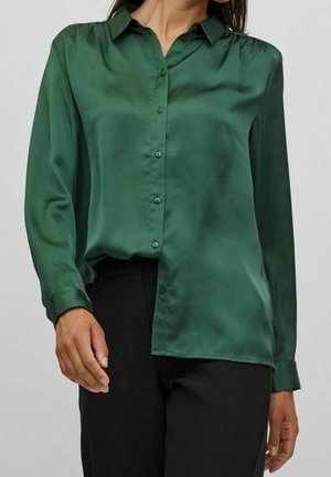 Femme portant un chemisier en satin vert forêt à manches longues avec des boutons sur le devant, rentré dans un pantalon noir.