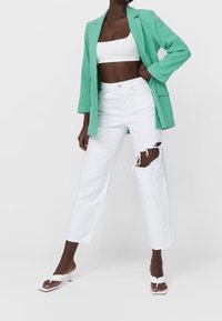 Muntgroene blazer over een wit crop-top, gecombineerd met versleten witte jeans. De schoeisel bestaat uit witte sandalen met hakken en een vierkante neus.