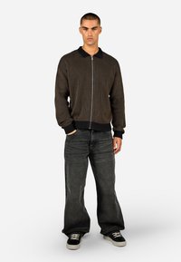 Rjava rebrasta jakna na zadrgo s črnim ovratnikom, v kombinaciji s širokimi temno sivimi jeans hlačami in črnimi čevlji z belimi podplati.