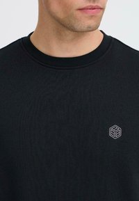 Sudadera negra con cuello redondo, textura acanalada y un pequeño logo geométrico bordado en el pecho.
