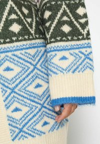 Maglione lavorato a maglia con motivi geometrici verdi, bianchi e blu, maniche a costine e una consistenza morbida. Principalmente crema con accenti blu.