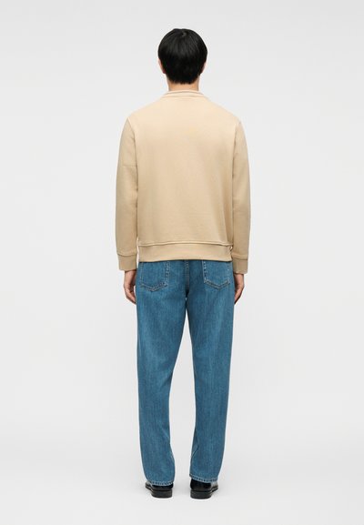 Άτομο που στέκεται με την πλάτη προς τα εμπρός, φορώντας μπεζ μπλούζα με μακριά μανίκια, μπλε τζιν και μαύρα παπούτσια, μπροστά σε ένα απλό ανοιχτόχρωμο φόντο.