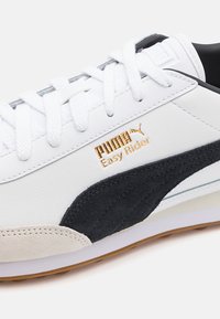 Zapatilla blanca de cuero con detalles en ante negro, perforaciones y logo dorado. Presenta cordones planos y una suela acolchada.