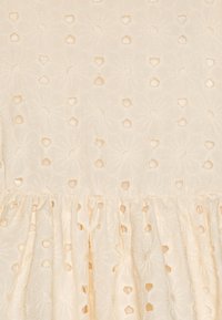 Tissu de couleur crème avec des motifs floraux brodés et des trous découpés. Présente un volan froncé le long du bord inférieur.