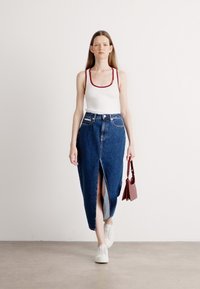 Jupe maxi en denim avec une fente à l'avant, associée à un débardeur blanc avec des bordures rouges. Le modèle porte des baskets blanches et tient un petit sac rouge.